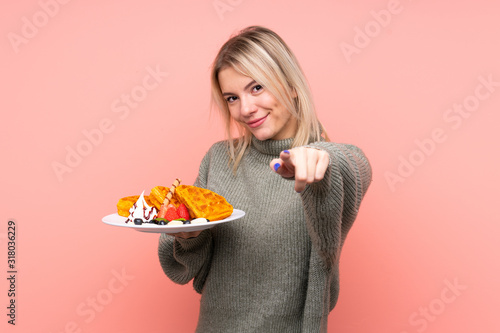 Young blonde woman holding waffles over isolated pink background points finger at you with a confident expression