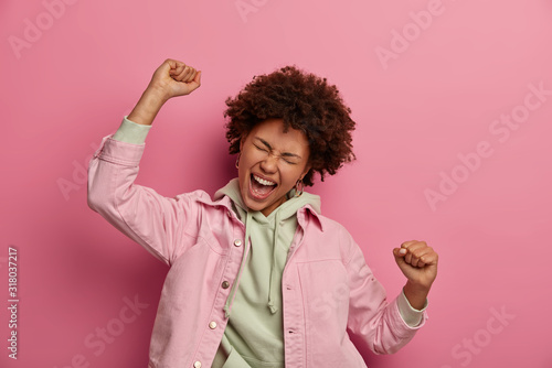 Overjoyed Afro American student makes victory dance, exclaims happily, being on cloud nine, achieves success, wears hoodie with jacket, closes eyes with pleasure, moves over rosy background.
