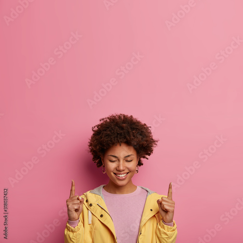 Positive Afro woman points above with closed eyes, feels glad to participate in advertising campaign, has toothy gentle smile, wears yelllow jacket, attracts attention upwards. Your promo content here