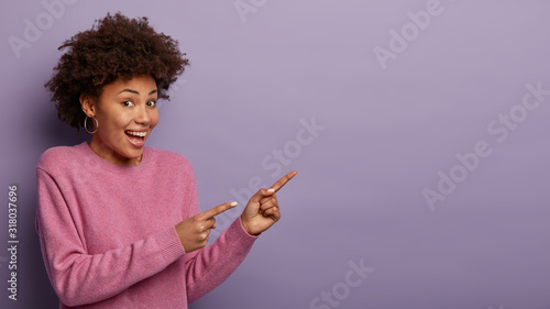 Look at this! Satisfied woman with Afro hairstyle, giggles from happiness, points two index fingers on blank space, wears casual jumper, shows blank space on purple wall for your promotional content