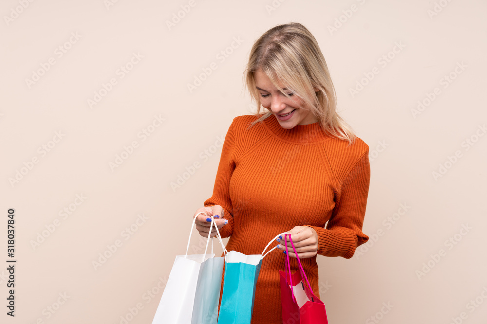 Young blonde woman over isolated background holding shopping bags and looking inside it Young blonde woman over isolated background holding shopping bags and looking inside it