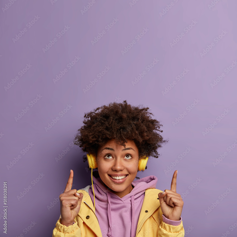 Φωτογραφία Glad positive woman has curly hairstyle, points both index finger on ceiling, shows copy space above, has good mood as listens amusing songs, wears stereo headphones on ear