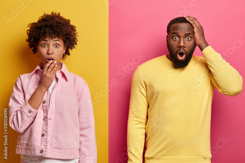 Shocked black female and male stare at camera, expresses great surprisement, open mouthes, hear unbelievable news, wear pastel pink and yellow clothes, stand together in studio. Oh no, how scary! photo