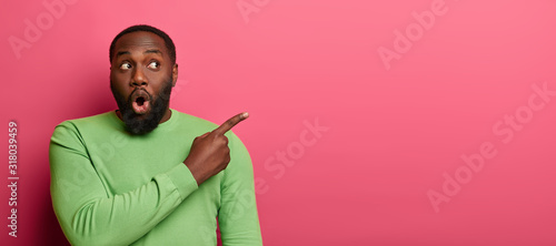Shocked emotive bearded man with black skin, wears bright green sweater, points at empty space, surprised by unexpected relevation, demonstrates place on pink wall for your promotional content