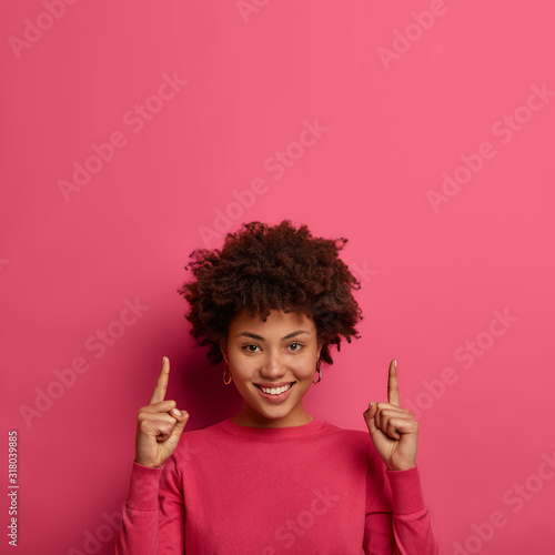 Glad woman points both fingers on copy space above, has friendly positive smile, wears bright clothes in one tone with background, suggests going upstairs, gives good advice what to buy on sale