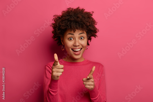 You are picked. Delighted cheerful friendly looking hipster girl has Afro hairstyle, makes finger gun gesture, indicates at camera happily, chooses someone in her team, wears casual bright wear