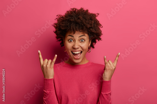 Positive curly haired girl makes horn gesture, enjoys rock n roll music, laughs with joy, wears casual jumper, feels upbeat and cool, isolated over bright pink background. Body language concept