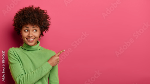 Go right there. Joyful friendly woman shows direction to shop, explains way, points fore finger on empty space, wears green jumper, poses against pink wall with copy place for advertising content photo