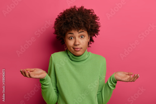 Indifferent confused black woman spreads palms confusingly, has doubts, wears green turtleneck, has hesitant face expression, isolated over pink background, has no idea, puzzled to give answer