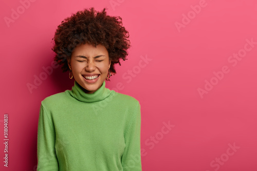Indoor shot of overemotive Afro American woman laughs at funny joke, closes eyes from joy, has overjoyed face expression, doesnt think about troubles, isolated over rosy wall with blank space © wayhome.studio 