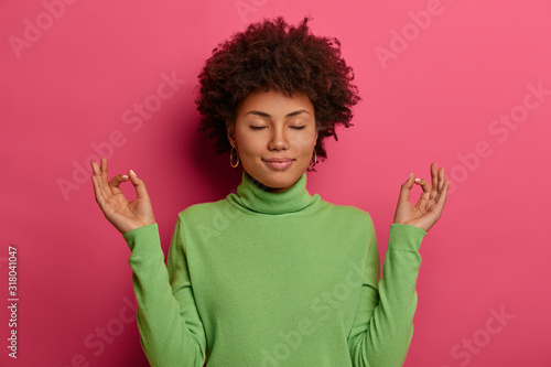 Relieved woman reaches nirvana, stands in lotus pose, meditates after work, closes eyes and breathes deeply, tries to sooth stress, practices yoga indoor, wears green casual jumper, isolated on pink
