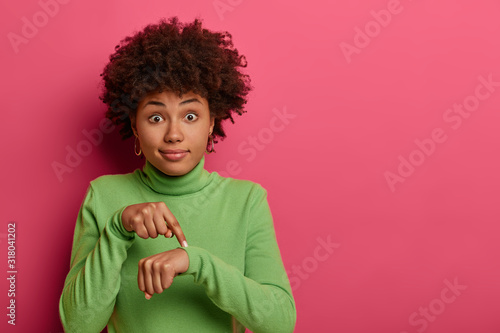 Time to go, hurry up! Shocked ethnic woman points at wrist, makes time gesture, shows we should do everything quickly, dressed in green clothes, stands against pink background. Its too late. photo