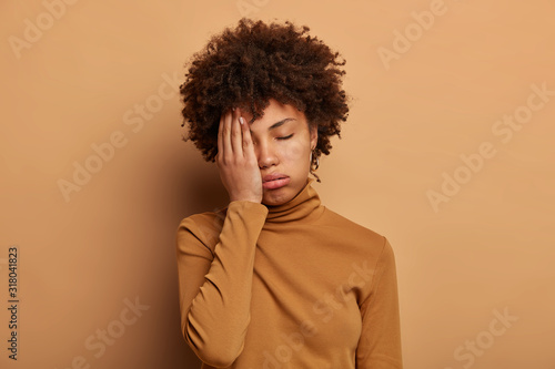 Photo of tired curly woman covers face with palm, feels overworked and fatigue, wants to sleep, tilts head, wears casual turtleneck, isolated over brown background. Tiredness, dissatisfaction concept