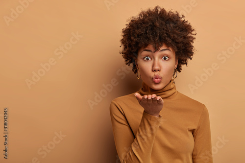 Passionate good looking woman with Afro hairstyle blows airkiss, holds palm near folded lips, makes flity message to boyfriend, stands indoor against brown background. Romantic feeling, tenderness © wayhome.studio 