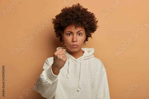 Serious angry ethnic woman threatens you with clenched fist, says I will show you how to disobey, wears casual white hoodie, promises to punish, looks with annoyance, isolated on beige background