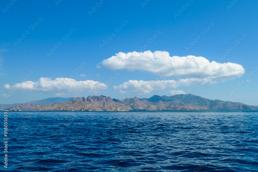 A view on idyllic island in Komodo National Park, Indonesia. There are few clouds above the island. Calm and clear surface of the sea. Island hoping. Perfect day for sailing A view on idyllic island in Komodo National Park, Indonesia. There are few clouds above the island. Calm and clear surface of the sea. Island hoping. Perfect day for sailing