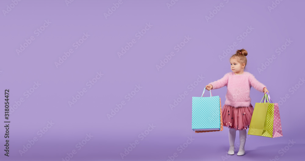 Portrait of a happy little child girl standing on a purple background holding shopping bags, package. Looking at the camera.