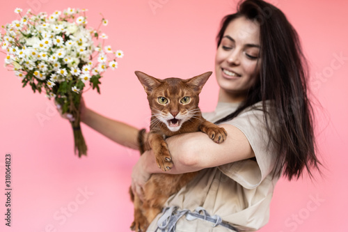Girl in a dress with a Bengal cat and bouqet of flowers on a pink background in front of camera
