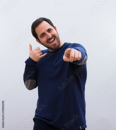 young handsome man smiling cheerfully and pointing to camera while making a call you later gesture, talking on phone against white wal.