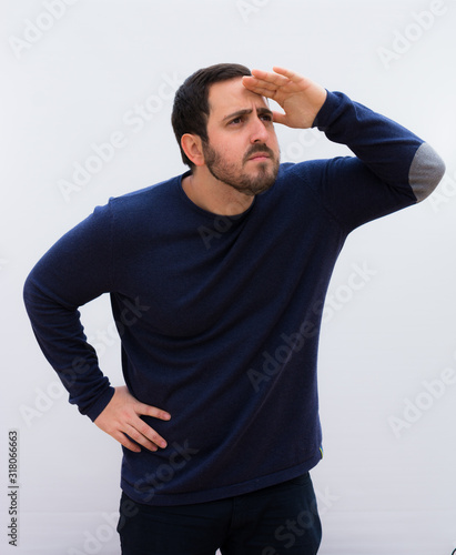 young handsome man looking bewildered and astonished, with hand over forehead looking far away, watching or searching against white wall.