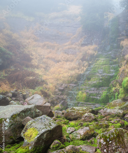 Scenic mountain forest waterfall, red rocky cliff covered by sunlit green moss and thick morning mist