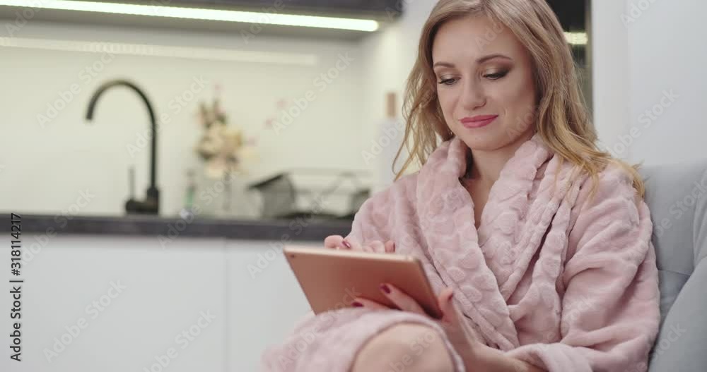 Portrait Beautiful female 30s in bathrobe using tablet pc, sitting on the sofa, looking at camera. Woman has make-up on face. Dolly shot Portrait Beautiful female 30s in bathrobe using tablet pc, sitting on the sofa, looking at camera. Woman has make-up on face. Dolly shot