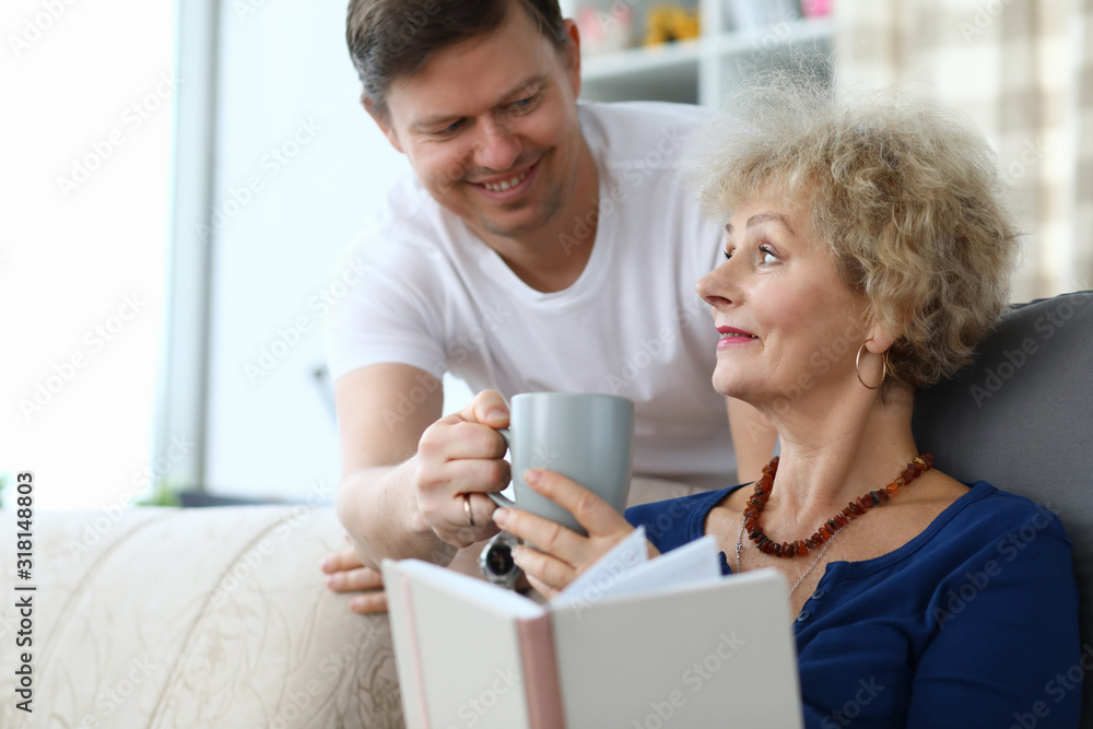 Portrait of smiling man giving cup of tea to mom. Woman sitting on sofa and reading book. Family spending time together. Warm relationship concept