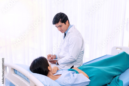 Hospitalized young woman lying in bed while doctor checking her healthcare. Friendly man physician examining female patient in hospital room ward while holding clipboard report in his hand.