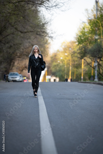 A young beautiful blonde girl in a black leather jacket is walking along an empty city road, the lower angle of the shooting