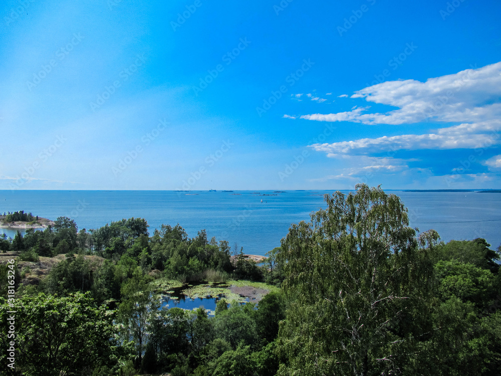 View from a hill with open sea, a small pond, trees, sailboat and some white clouds on a sunny day.