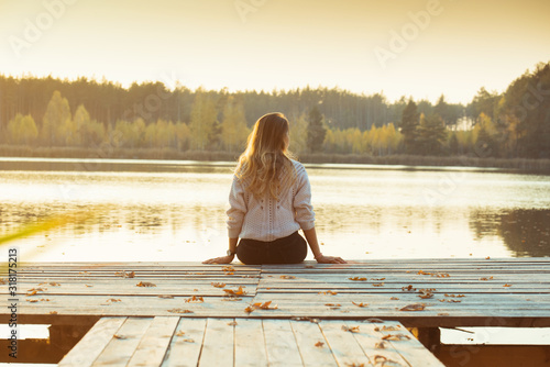 A young slender blonde sits on a wooden pier with her back . photo