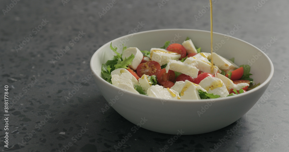add olive oil to salad with mozzarella, cherry tomatoes and frisee leaves in white bowl on terrazzo surface add olive oil to salad with mozzarella, cherry tomatoes and frisee leaves in white bowl on terrazzo surface