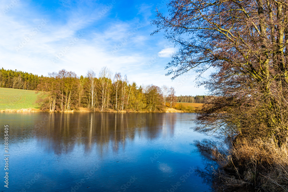 Spring lake in czech countryside. Blue sky, green field and frozen water.