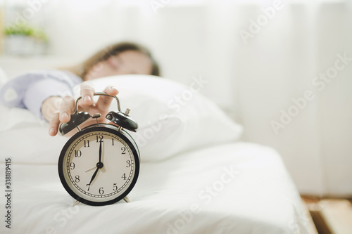 young asian woman sleeping on bed pressing snooze button on black vintage alarm clock at seven o'clock morning in bed room at home, lifestyle, good morning, healthy sleep and joyful weekend concept