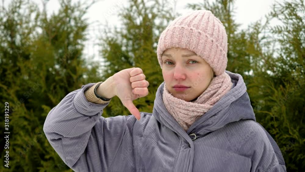 Young beautiful woman standing on green fur trees background, expressing discontent and showing thumb down gesture at camera. Portrait of girl with sign of dislike. 4K Young beautiful woman standing on green fur trees background, expressing discontent and showing thumb down gesture at camera. Portrait of girl with sign of dislike. 4K