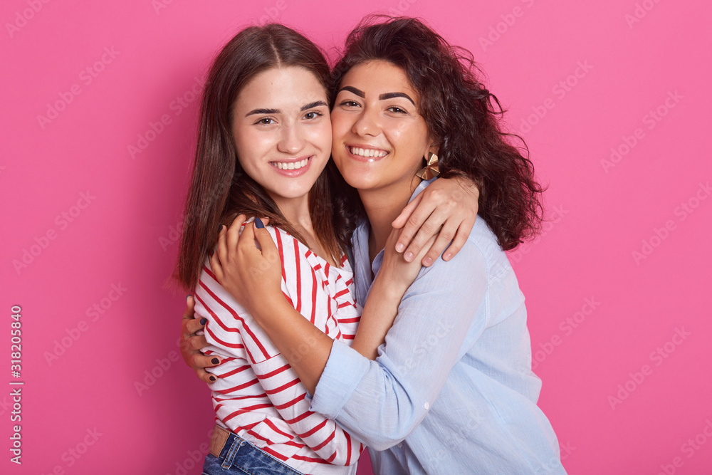 Horizontal shot of beautiful young friends with dark hair, looking smiling diretly at camera. Lady with wavy hair wears blue shirt, girl with staright hairstyle dresses casual shirt with red stripes. Horizontal shot of beautiful young friends with dark hair, looking smiling diretly at camera. Lady with wavy hair wears blue shirt, girl with staright hairstyle dresses casual shirt with red stripes.