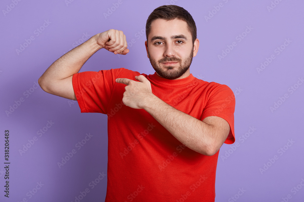 Close up portrait of happy strong man dresses casual red t shirt showing his biceps on arm and pointing with index finger, male looking at camera while standing isolated over purple background. Close up portrait of happy strong man dresses casual red t shirt showing his biceps on arm and pointing with index finger, male looking at camera while standing isolated over purple background.