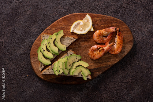 Wholegrain toast bread slices with guacamole, fried shrimp and fried bacon pieces on wooden board Selective Focus, Focus on the front of the shrimp on the first bread