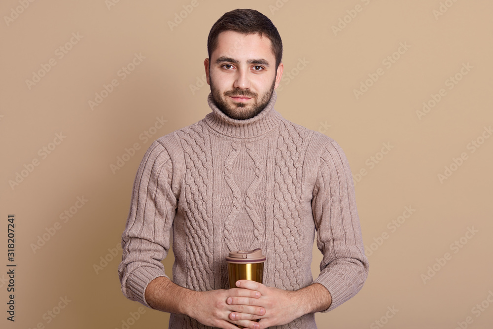 Studio shot of young serious Caucasian man holding coffee cup isolated over beige background, handsome male having thermo mug in hands, enjoying hot beverage, looking at camera, looks confident. Studio shot of young serious Caucasian man holding coffee cup isolated over beige background, handsome male having thermo mug in hands, enjoying hot beverage, looking at camera, looks confident.