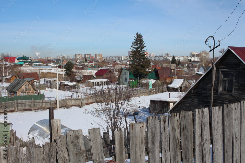 wooden summer cottages in winter in a garden community in the snow near a modern city with a fence in the village