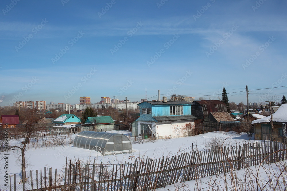 wooden summer cottages in winter in a garden community in the snow near a modern city with a fence in the village