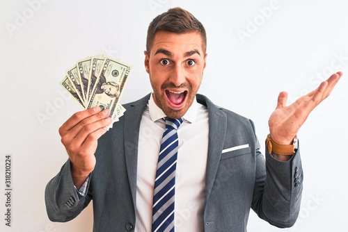 Young handsome business man holding bunch of dollars banknotes over isolated background very happy and excited, winner expression celebrating victory screaming with big smile and raised hands