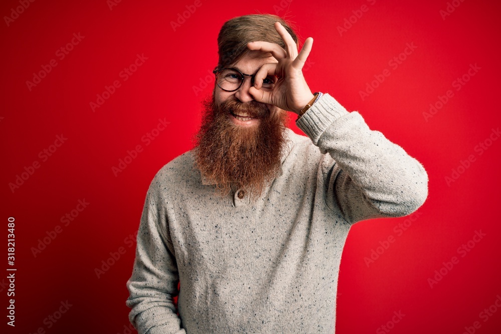 Handsome Irish redhead man with beard wearing casual sweater and glasses over red background doing ok gesture with hand smiling, eye looking through fingers with happy face. Handsome Irish redhead man with beard wearing casual sweater and glasses over red background doing ok gesture with hand smiling, eye looking through fingers with happy face.