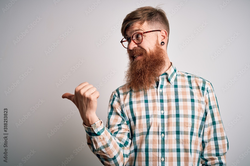 Handsome Irish redhead man with beard wearing glasses and hipster shirt smiling with happy face looking and pointing to the side with thumb up. Handsome Irish redhead man with beard wearing glasses and hipster shirt smiling with happy face looking and pointing to the side with thumb up.
