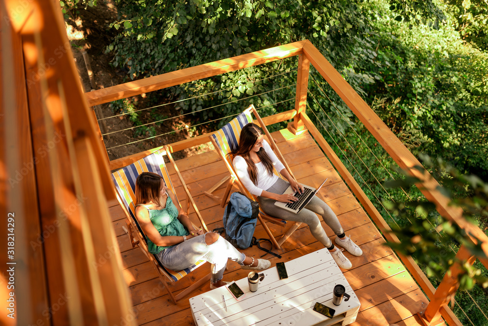 High angle image of two young women enjoying on a wooden cabin terrace in forest. - obrazy, fototapety, plakaty 
