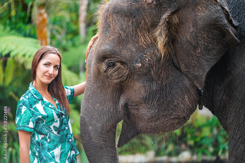 Fototapeta premium A Caucasian woman in a bright shirt poses next to an elephant. Close up. Jungle forest on the background