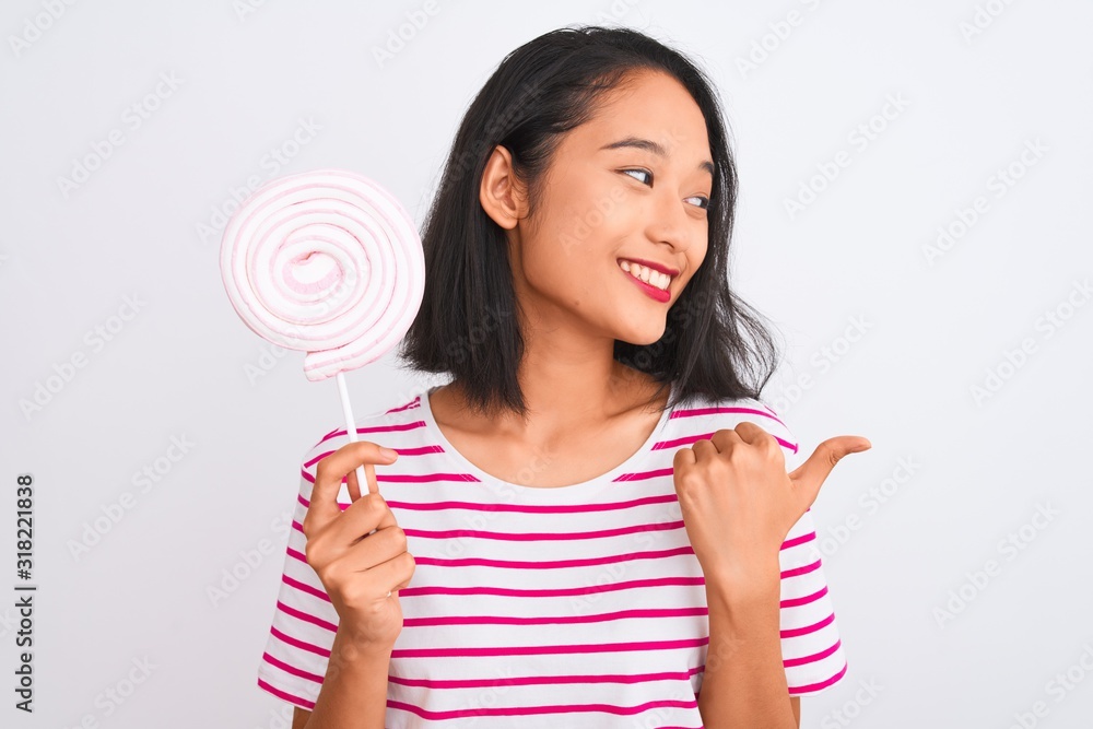Young beautiful chinese woman eating lollipop standing over isolated white background pointing and showing with thumb up to the side with happy face smiling Young beautiful chinese woman eating lollipop standing over isolated white background pointing and showing with thumb up to the side with happy face smiling
