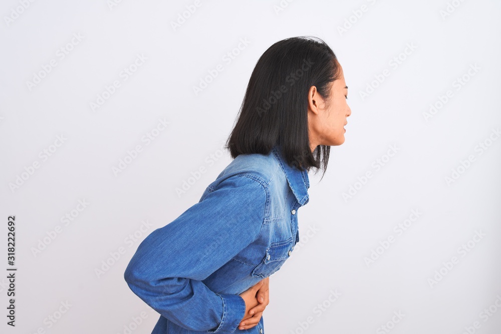 Young beautiful chinese woman wearing denim shirt standing over isolated white background with hand on stomach because indigestion, painful illness feeling unwell. Ache concept. Young beautiful chinese woman wearing denim shirt standing over isolated white background with hand on stomach because indigestion, painful illness feeling unwell. Ache concept.