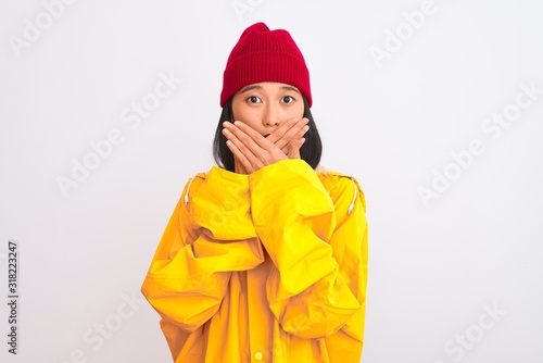 Young beautiful chinese woman wearing raincoat and wool cap over isolated white background shocked covering mouth with hands for mistake. Secret concept.