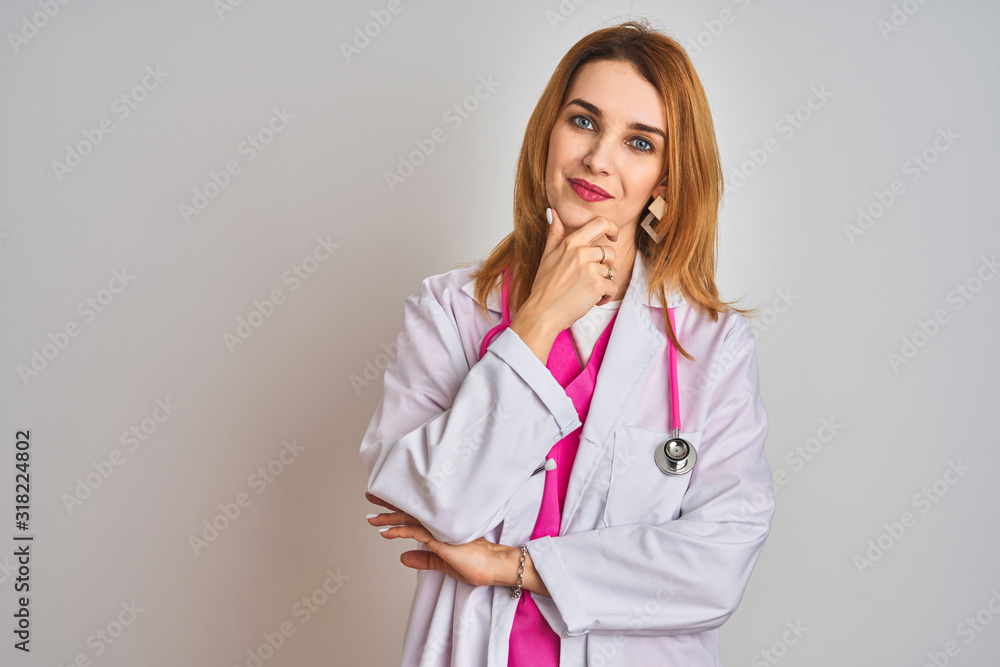 Redhead caucasian doctor woman wearing pink stethoscope over isolated background looking confident at the camera smiling with crossed arms and hand raised on chin. Thinking positive. Redhead caucasian doctor woman wearing pink stethoscope over isolated background looking confident at the camera smiling with crossed arms and hand raised on chin. Thinking positive.